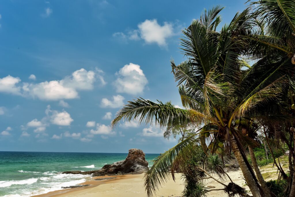 A tropical beach in Malaysia with golden sand, palm trees, turquoise waters, and a rocky formation under a bright blue sky.